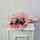 A bouquet of pink roses and white baby's breath flowers wrapped in pink tissue paper with gold ribbon trim, resting on a white cabinet against a light gray wall.