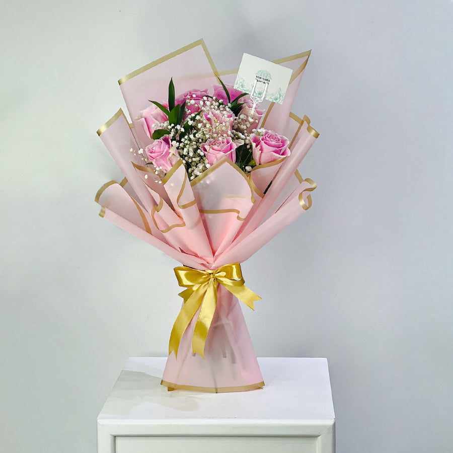 A bouquet of pink roses wrapped in light pink paper with gold trim and tied with a gold ribbon, displayed on a white pedestal against a light gray background.