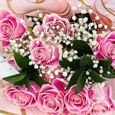 A close-up of a flower bouquet featuring vibrant pink roses mixed with delicate white baby's breath and dark green leaves, wrapped in pink ribbon and paper.