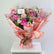 A vibrant bouquet of pink and purple flowers wrapped in pink paper with gold trim, sitting on a white table against a light gray background.