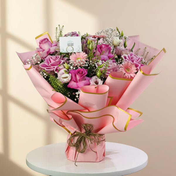 A vibrant bouquet of pink and purple flowers wrapped in pink paper with gold trim, sitting on a white table against a light gray background.