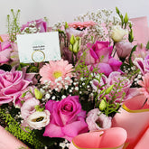 A vibrant floral bouquet filled with pink and light pink flowers, including roses, gerbera daisies, and lisianthus, accented with green foliage and small white baby's breath. A white card with the text 'VIVA FLORA ماري لومبي' is tucked into the arrangement, set against a soft pink and white background.