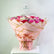 A large bouquet of pink, red, and white flowers wrapped in light pink paper with gold geometric lines, tied with a pink ribbon, displayed on a white pedestal against a white wall.