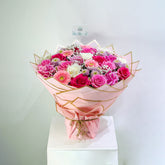 A vibrant bouquet of pink, magenta, and white roses with gerbera daisies, wrapped in pink paper with gold geometric borders and tied with a pink ribbon, displayed on a white pedestal against a light gray background.