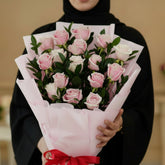 A bouquet of pink and white roses wrapped in light pink tissue paper with a red ribbon tied in a bow at the base, set against a light marble-patterned background.