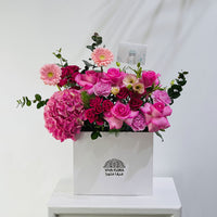 A vibrant pink floral arrangement featuring roses, hydrangeas, and daisies in a white square box with the brand name 'VIVA FLORA' printed on the front, displayed on a white pedestal against a white wall.