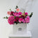 A vibrant pink floral arrangement featuring roses, hydrangeas, and daisies in a white square box with the brand name 'VIVA FLORA' printed on the front, displayed on a white pedestal against a white wall.