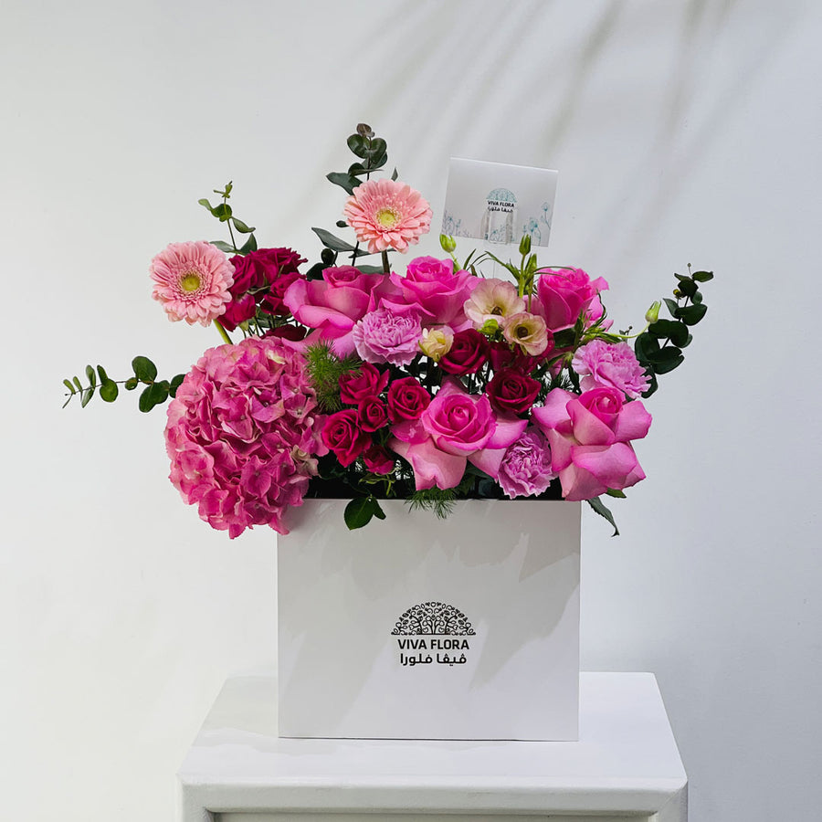 A vibrant pink floral arrangement featuring roses, hydrangeas, and daisies in a white square box with the brand name 'VIVA FLORA' printed on the front, displayed on a white pedestal against a white wall.