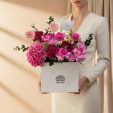 A vibrant pink floral arrangement featuring roses, hydrangeas, and daisies in a white square box with the brand name 'VIVA FLORA' printed on the front, displayed on a white pedestal against a white wall.