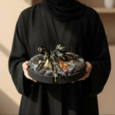 A round black gift box filled with individually wrapped chocolates, tied with a black ribbon printed with gold lettering, displayed on a beige pedestal against a warm beige background.
