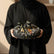A round black gift box filled with individually wrapped chocolates, tied with a black ribbon printed with gold lettering, displayed on a beige pedestal against a warm beige background.