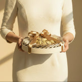 A round white gift box filled with individually wrapped chocolates or candy, tied with a large satin ribbon, displayed on a matching white pedestal against a warm beige background.