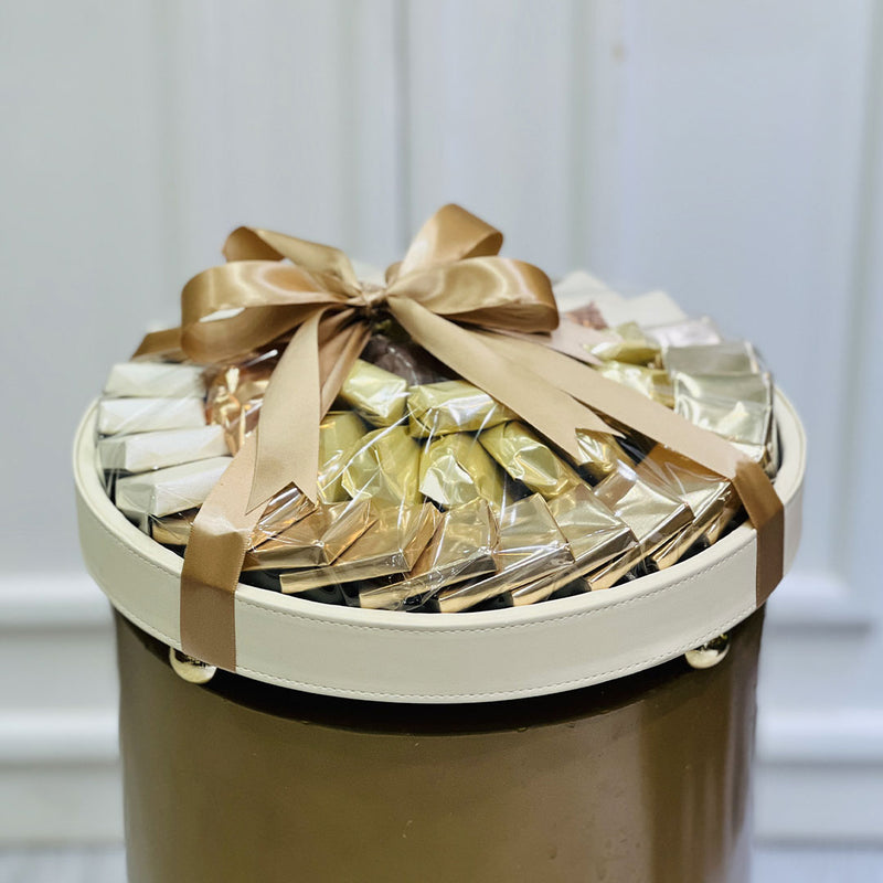 Round gift basket with gold ribbons filled with wrapped chocolate on a neutral background