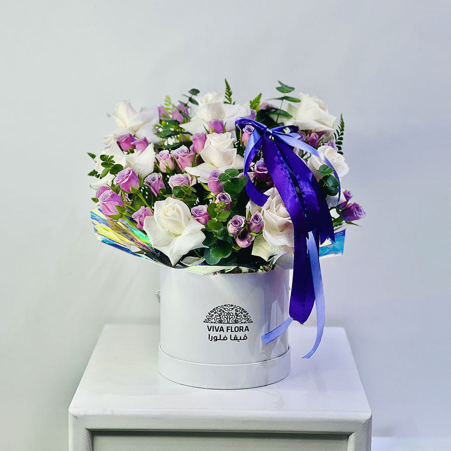 A floral arrangement of white and light purple roses with green foliage, presented in a round white box with the brand name VIVA FLORA, tied with a purple ribbon, set on a white pedestal against a light gray background.