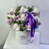 A round white gift box filled with a bouquet of white and light purple roses, green foliage, and a large purple ribbon bow, displayed on a white marble-like surface against a light gray wall. The box is branded VIVA FLORA.