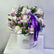 A round white gift box filled with a bouquet of white and light purple roses, green foliage, and a large purple ribbon bow, displayed on a white marble-like surface against a light gray wall. The box is branded VIVA FLORA.