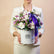 A floral arrangement of white and light purple roses with green foliage, presented in a round white box with the brand name VIVA FLORA, tied with a purple ribbon, set on a white pedestal against a light gray background.