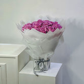 A large bouquet of vibrant pink roses wrapped in white tissue paper and tied with a gray ribbon, displayed on a white pedestal against a plain light gray wall.
