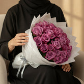 A large bouquet of vibrant pink roses wrapped in white tissue paper and tied with a gray ribbon, displayed on a white pedestal against a plain light gray wall.