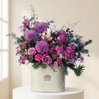 A vibrant floral arrangement in a white cylindrical box, featuring prominent purple and pink flowers with green foliage, displayed on a white pedestal against a white studio backdrop.