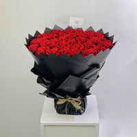A large bouquet of red roses wrapped in black paper and tied with twine, displayed on a white pedestal against a light gray background.