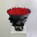 A large bouquet of red roses wrapped in black paper and tied with twine, displayed on a white pedestal against a light gray background.