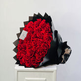 A large bouquet of red roses wrapped in black paper, displayed on a white pedestal against a plain light gray wall.