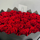 A heart-shaped arrangement of vibrant red roses in a dark gray paper-lined box, with a small white card inserted in the center.