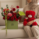 A red plush teddy bear sits on a white pedestal next to a green box of red and pink flowers with green foliage, set against a light beige wall with soft window shadows.