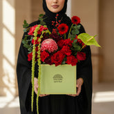 A vibrant floral arrangement featuring red roses, red anthurium, a pink chrysanthemum, and green foliage, presented in a light green square box with the brand name 'VIVA FLORIDA' printed on the front, set on a white pedestal against a beige wall with soft window shadows.