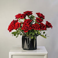 A bouquet of 12 red roses with green leaves arranged in a black faceted geometric vase, sitting on a white marble-topped table against a neutral gray background.