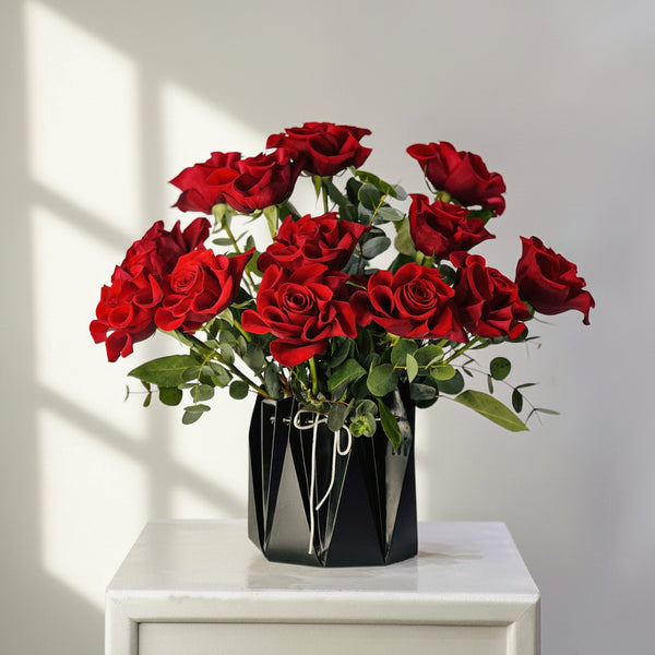 A bouquet of 12 red roses with green leaves arranged in a black faceted geometric vase, sitting on a white marble-topped table against a neutral gray background.