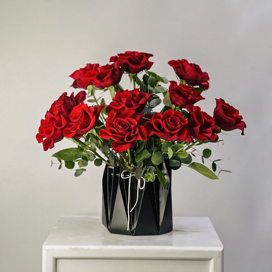 A bouquet of 12 red roses with green leaves arranged in a black faceted geometric vase, sitting on a white marble-topped table against a neutral gray background.