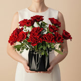 A bouquet of vibrant red roses with green leaves arranged in a black geometric faceted vase, placed on a white marble-topped table against a light gray wall with soft window shadow patterns.