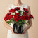 A bouquet of vibrant red roses with green leaves arranged in a black geometric faceted vase, placed on a white marble-topped table against a light gray wall with soft window shadow patterns.