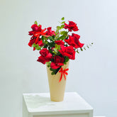 A bouquet of Fresh red roses with green leaves arranged in a tall cream-colored vase tied with a red ribbon, displayed on a white pedestal against a light gray background.