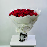 A large bouquet of red roses wrapped in white paper and tied with a black ribbon, displayed on a white pedestal against a light gray background.