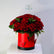 A large bouquet of red roses with green leaves arranged in a bright red cylindrical box with butterfly cutouts and a black base, displayed on a white surface against a light gray background.