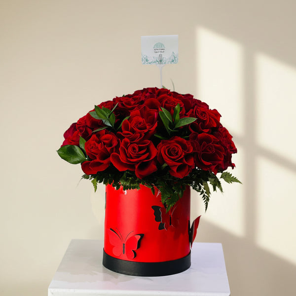 A large bouquet of red roses with green leaves arranged in a bright red cylindrical box with butterfly cutouts and a black base, displayed on a white surface against a light gray background.
