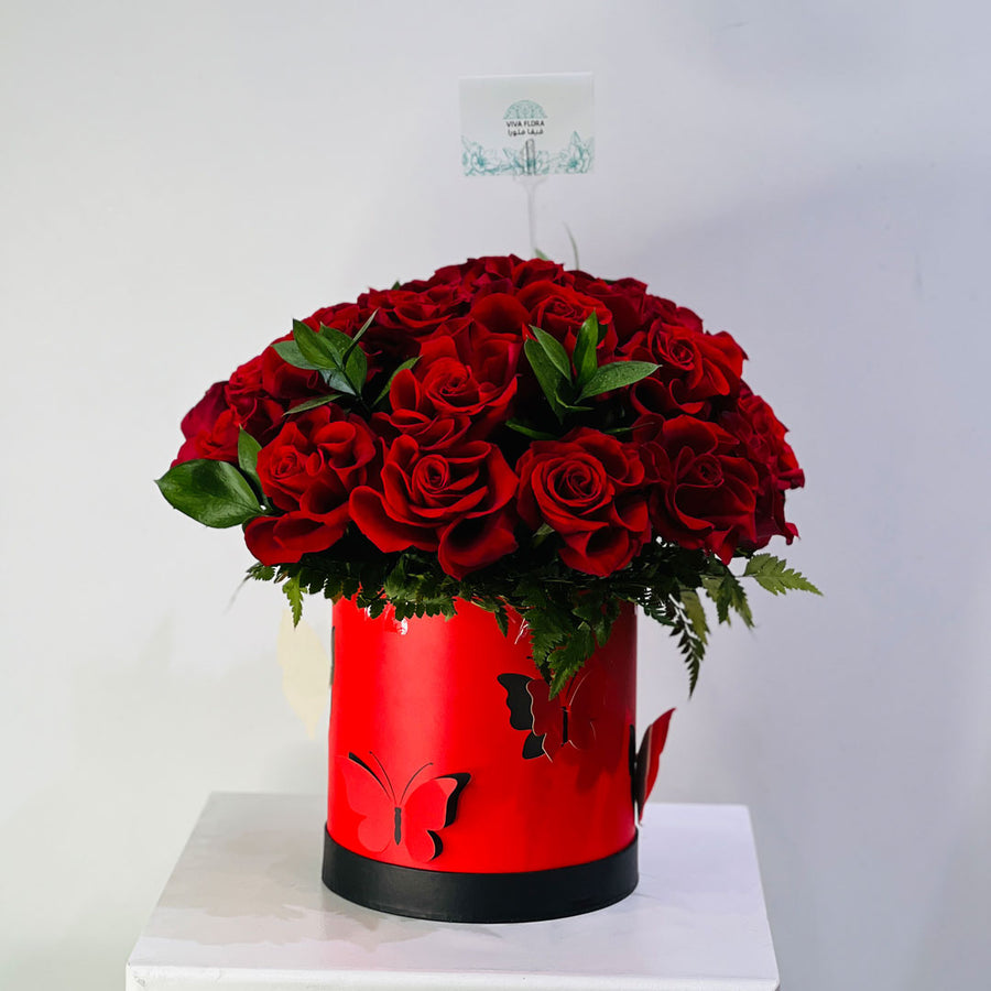 A large bouquet of red roses with green leaves arranged in a bright red cylindrical box with butterfly cutouts and a black base, displayed on a white surface against a light gray background.