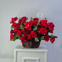 A large arrangement of vibrant red roses and green foliage in a brown wicker basket, displayed on a white cabinet against a light gray wall.