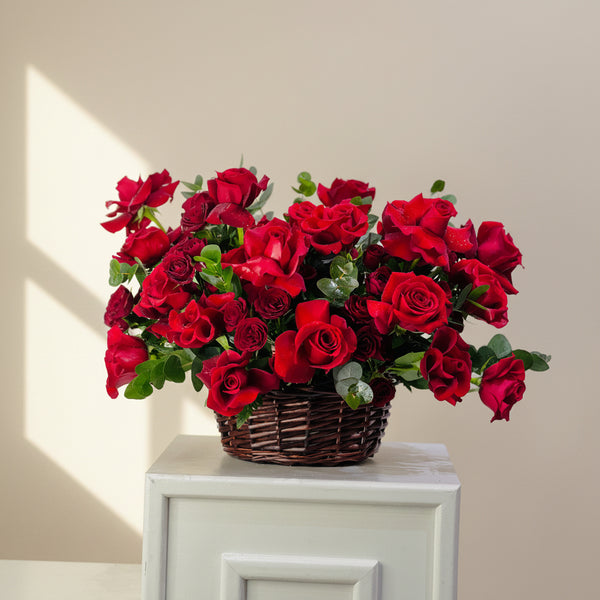 A large arrangement of vibrant red roses and green foliage in a brown wicker basket, displayed on a white cabinet against a light gray wall.
