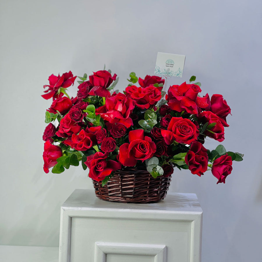 A large arrangement of vibrant red roses and green foliage in a brown wicker basket, displayed on a white cabinet against a light gray wall.