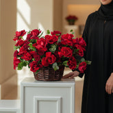 A large arrangement of vibrant red roses and green foliage in a brown wicker basket, displayed on a white cabinet against a light gray wall.