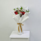 A bouquet of red roses mixed with white baby's breath and green eucalyptus stems, wrapped in white paper with a gold ribbon, displayed on a white pedestal against a light gray background.