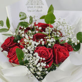 A bouquet of red roses mixed with white baby's breath and green eucalyptus leaves, wrapped in white paper with a card that reads 'VIVA FLORA'.
