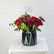 A bouquet of deep red roses and green eucalyptus stems arranged in a black faceted geometric vase, tied with a white string, displayed on a white table against a light gray background.