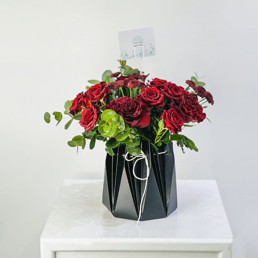 A bouquet of deep red roses and green eucalyptus stems arranged in a black faceted geometric vase, tied with a white string, displayed on a white table against a light gray background.
