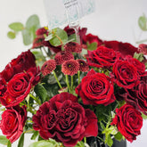 A close-up of a flower bouquet featuring deep red roses and small burgundy daisy-like blooms with green leaves, photographed against a clean white background. A hanging tag is partially visible at the top.