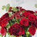A close-up of a flower bouquet featuring deep red roses and small burgundy daisy-like blooms with green leaves, photographed against a clean white background. A hanging tag is partially visible at the top.